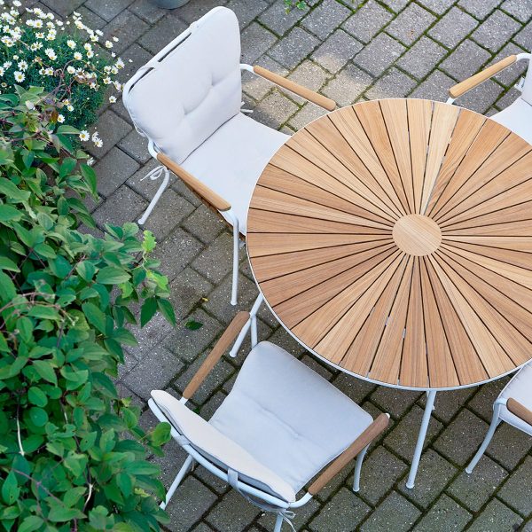 Et fugleperspektiv af et MANDALAY - Marguerit Helhynde rundt terrassebord i træ med sunburst-design, ledsaget af to hvide stole, på en murstensoverflade ved siden af grønne planter.
