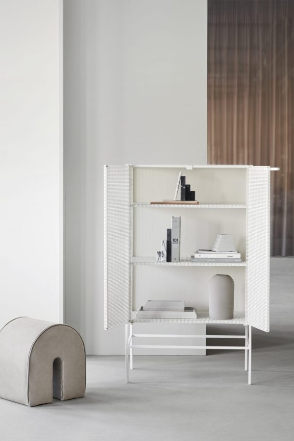 Et moderne, minimalistisk værelse med en hvid perforeret foldeskærm, der er åben omkring en elegant reol fyldt med bøger og dekorative genstande. Et gråt, bueformet møblersæde er placeret til venstre.