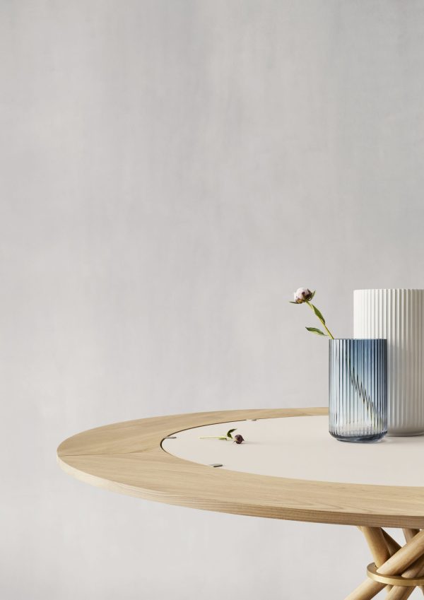 Et minimalistisk setup med et rundt, lyst træbord med en tyndstilket lilla blomst i en tekstureret blå glasvase, mod en blød grå baggrund, er ideel til at fremvise designmøbler.