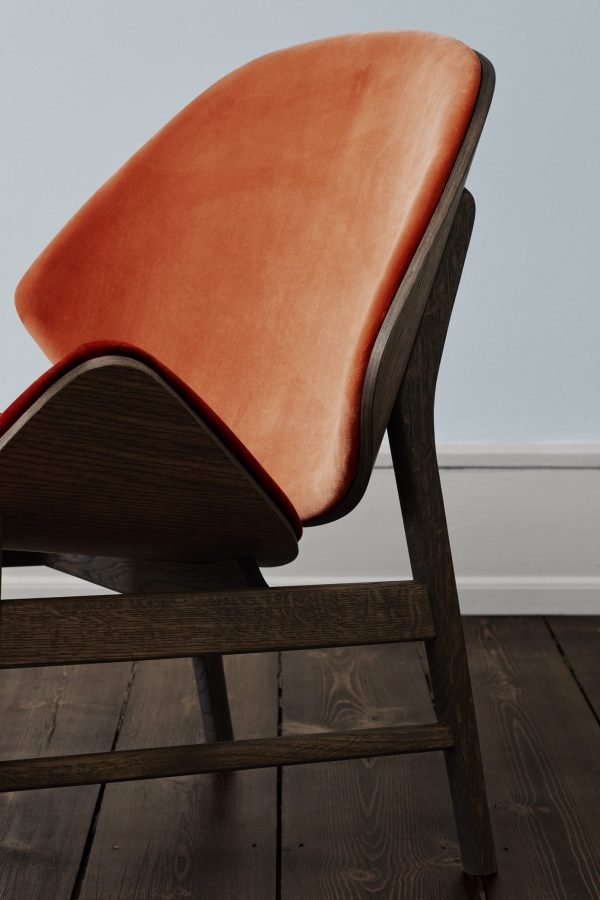 Set fra siden af en moderne stol med et slankt træstel og en glat WARM NORDIC - The Orange Loungestol-pude, placeret mod en simpel hvid væg på et mørkt trægulv.