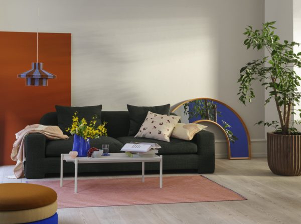 En moderne stue med en mørkegrøn sofa prydet med puder, et lille hvidt sofabord med en bog og gule blomster, en stor potteplante og farverige indretningselementer, herunder et orange tæppe og en blå bueformet dekoration af TomorrowsDesign.
