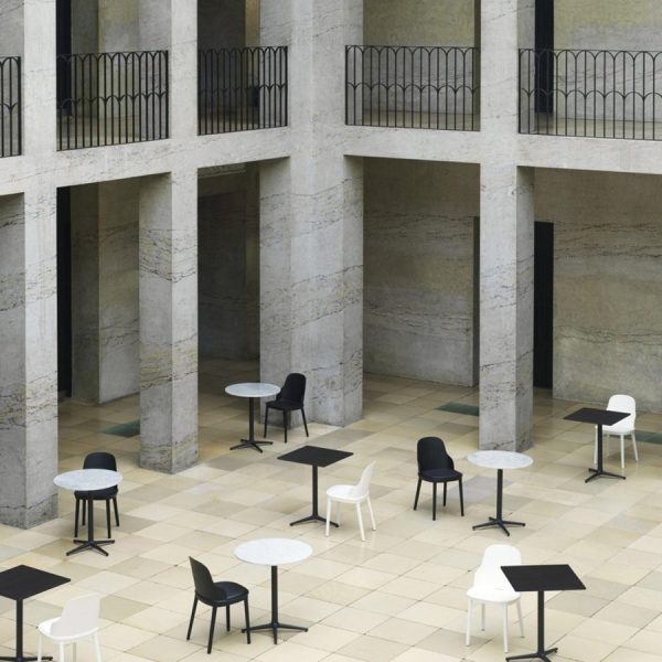 Et arkitektonisk indendørs rum med store betonsøjler og to niveauer af altaner. Gulvet er oversået med sorte og hvide design borde og stole. Vægge og gulve viser teksturerede stenoverflader.