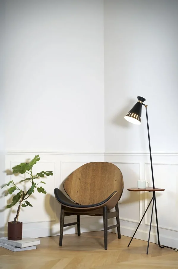 Et minimalistisk værelseshjørne med en træstol med buet ryglæn, en stående lampe vinklet mod stolen og et lille sidebord med en plante. Væggene har hvide lister, og der er et beige klinkegulv med accent af stilfulde designmøbler.