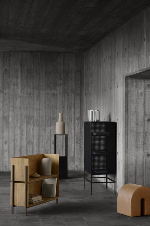Et minimalistisk værelse med grå trævægge og -loft, med en trælænestol, en sort reol og to moderne, skulpturelle stykker indretning. Interiøret har fokus på rene linjer og funktionel enkelhed.