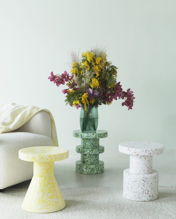 En farverig buket blomster i en grøn glasvase på en lysegrøn baggrund, flankeret af tre NORMANN COPENHAGEN - Bit Skammel Cone taburetter.