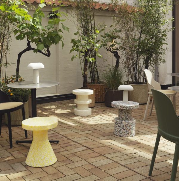 Et roligt udendørs terrasseområde med et udvalg af unikke NORMANN COPENHAGEN - Bit Skammel Cone taburetter og et bord, oplyst af en bordlampe. omgivelserne har murstensgulve og er omgivet af frodige planter og træer mod en hvid mur.