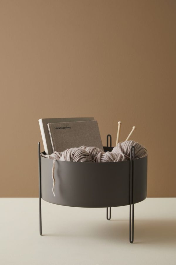 En sort WOUD - Pidestall Plantekasse Mellem indeholdende tre garnnøgler i grå og beige nuancer, med strikkepinde og en bog med titlen "livsrytmer" på beige baggrund.