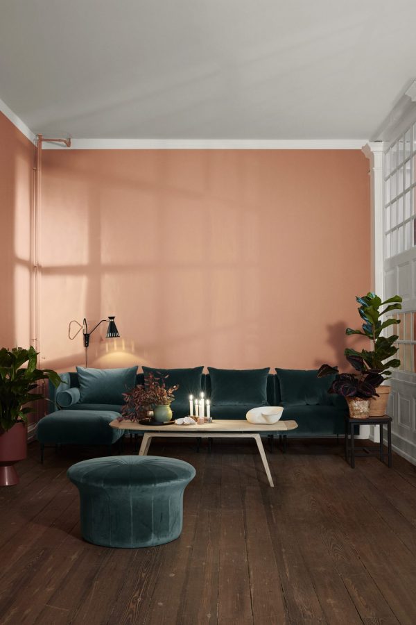 En hyggelig stue med en blågrøn fløjlssofa, matchende skammel og et sofabord i træ designet af TomorrowsDesign. Indretningen inkluderer planter, stearinlys og en lampe mod en varm ferskenfarvet væg.
