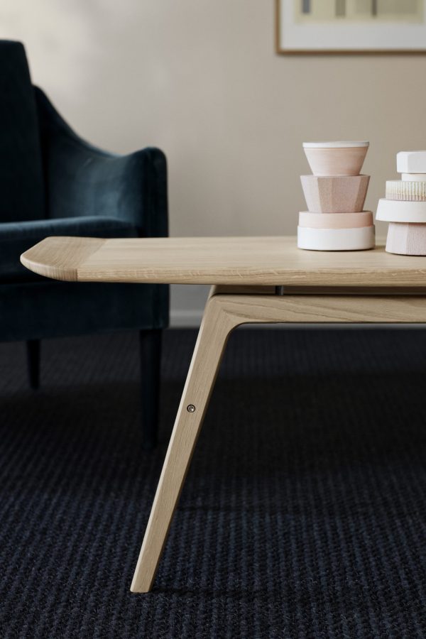 Et træbord med en glat finish og tilspidsede ben sidder i et rum med et mørkt tæppe og en grøn polstret stol. På toppen af bordet er der pænt stablet runde beholdere, der viser TomorrowsDesign. Et indrammet billede hænger på væggen i baggrunden.