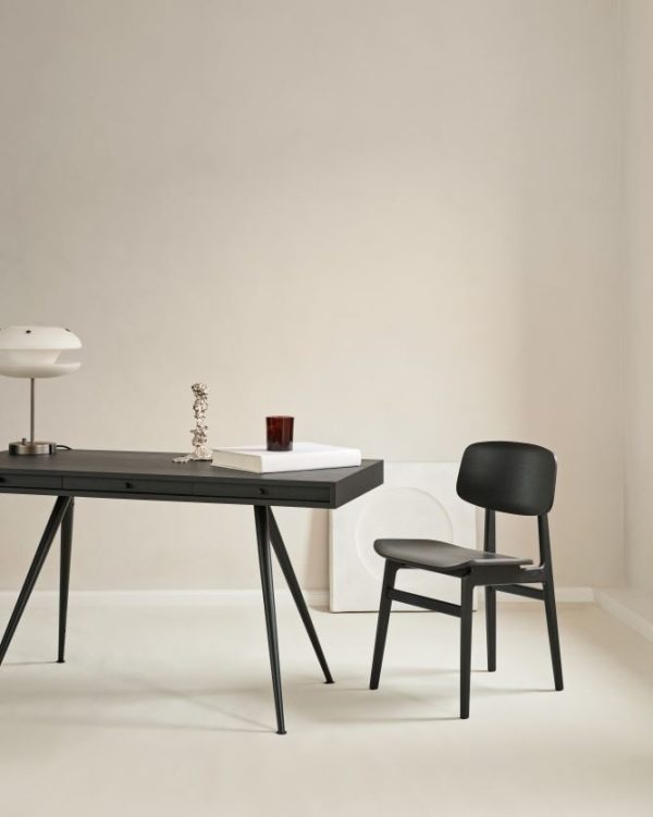 Et minimalistisk kontorlokale med et elegant sort skrivebord med en enkelt skuffe, parret med en NORR11 - NY11 Spisebordsstol. Skrivebordet rummer en lampe, en lille potteplante og et rødt glas. Lys beige vægge og gulv.