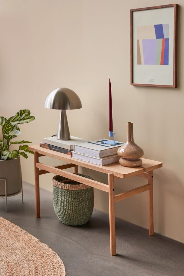 En pænt arrangeret interiørscene med en HÜBSCH - Align Bænk af træ med bøger, en metallisk lampe og dekorative vaser. et indrammet abstrakt kunstværk hænger ovenover, ved siden af en potteplante. et tekstureret tæppe er delvist synligt i bunden.