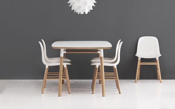 En moderne spisestue med et rektangulært bord med træben og en glat, lys bordplade, flankeret af tre designstole i skandinavisk stil med træben og hvide sæder, mod en grå væg. En hvid, skulpturel pendel hænger ovenover.