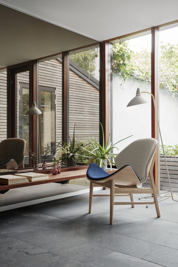 Moderne stue med store vinduer, et lækkert træbord, stilfulde stole og en gulvlampe. Rummet er udsmykket med potteplanter, der styrker forbindelsen til den udendørs terrasse og afspejler gennemtænkt indretning.