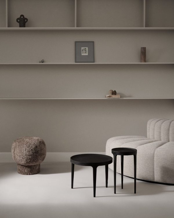 En minimalistisk stue med en tekstureret beige NORR11 - Ghost Sofabord, et rundt sort sofabord og en unik stenstol. Tomme hylder med et par små indretningsgenstande fremhæver det fredfyldte, neutrale rum.