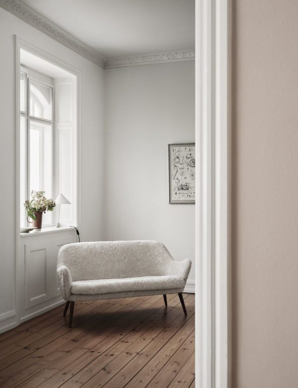 En minimalistisk stue med en hvid tekstureret sofa, trægulve og et stort vindue med en blomstervase på karmen. TomorrowsDesign væglister og en indrammet plakat tilføjer elegance til rummet.