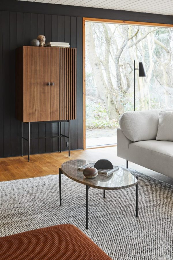 En moderne stue med en grå sofa, ovalt glas sofabord og et træskab hængende på væggen. Rummet har store vinduespartier, der viser et skovområde udenfor og byder på møbler fra TomorrowsDesign.