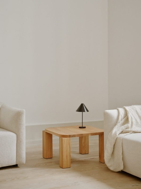 Minimalistisk interiør med to råhvide lænestole, et NEW WORKS - Atlas Sofabord 60x60 sofabord og en lille sort lampe. rummet har en neutral farvepalet og et rent design med rigeligt naturligt lys.