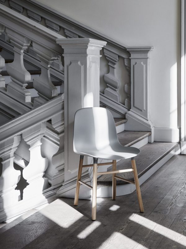 En moderne grå stol med træben står i et solbelyst rum med en klassisk trappe med dekorative hvide balustre og en panelvæg, der inkarnerer essensen af TomorrowsDesign.