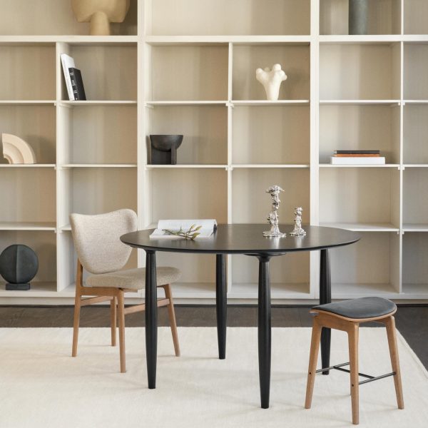 En moderne og minimalistisk spiseplads med et NORR11 Oku Spisebord Ø120 med to kontrasterende stole. bagved er en stor hvid reol med bøger, skulpturer og dekorative genstande.