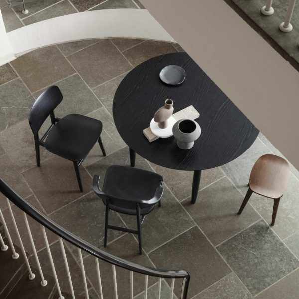Et billede ovenfra af en NORR11 - Oku Spisebord Ø120 med en tekstureret finish, ledsaget af fire moderne stole i forskellige stilarter og farver. bordet er dækket med minimalistisk keramisk keramik. omgivelserne har et klinkegulv og en del af et trappegelænder.