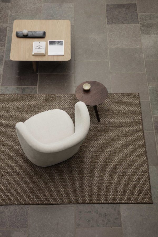 Moderne stueopsætning med designmøbler inklusive en tekstureret beige lænestol, et lille sofabord i træ med bøger og et rundt sidebord med en kop. Layoutet inkluderer et sisal tæppe på et klinkegulv.