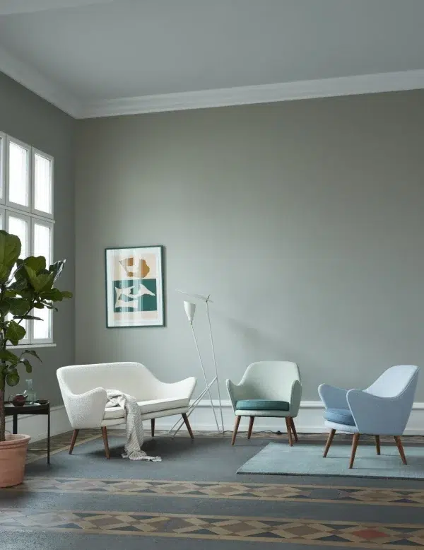 En minimalistisk stue med en blød grå væg, to vinduer med naturligt lys, en hvid sofa og tre pastelfarvede stole. En dekorativ lampe står ved siden af et indrammet abstrakt kunstværk. Et mønstret områdetæppe dækker gulvet. Interiørdesignet afspejler rene linjer og moderne enkelhed.