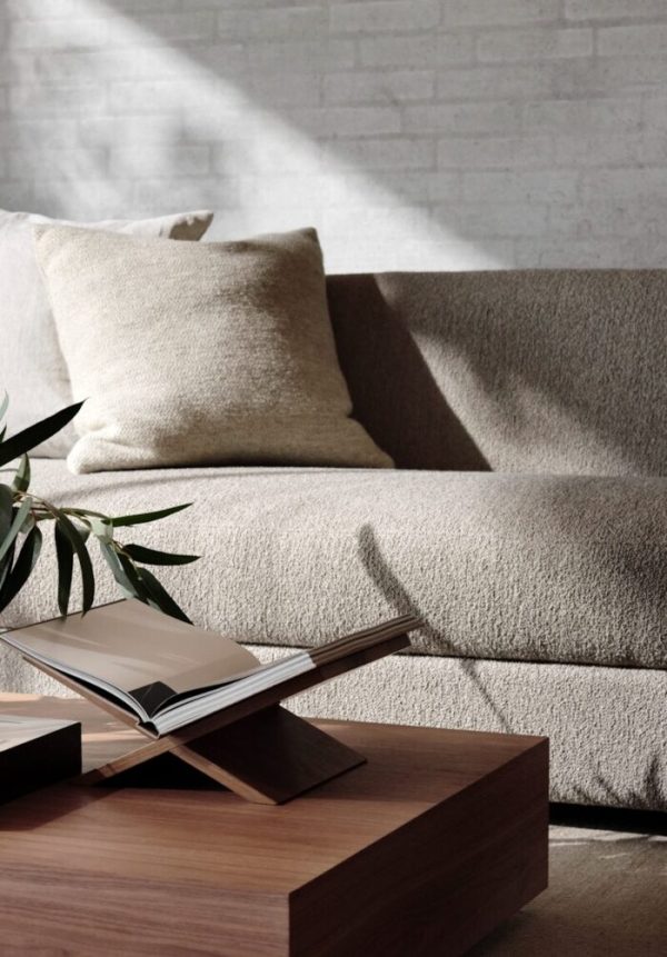 Et hyggeligt moderne opholdsrum med en beige sofa med hynder, et sofabord i træ med designmøbler-bøger på og en potteplante, sat op ad en hvid murstensvæg med sollys, der filtrerer igennem.