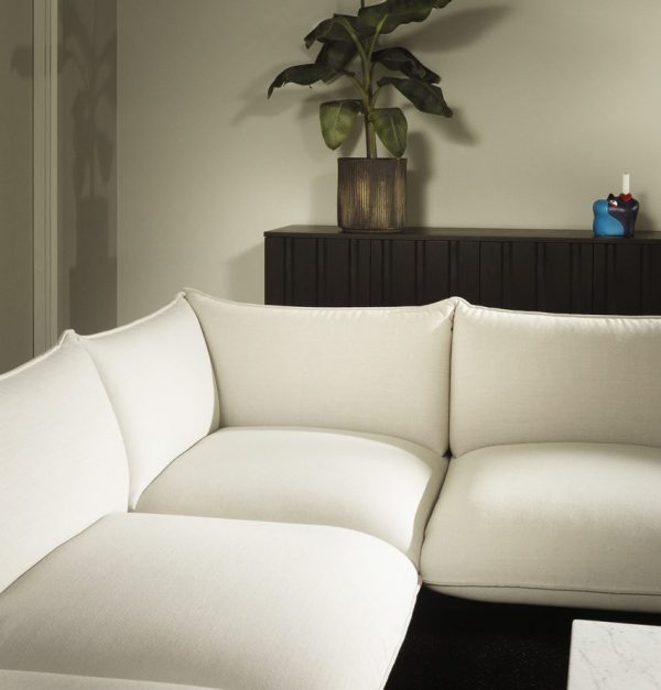 Et hjørne af en moderne stue med en beige NORMANN COPENHAGEN - Ark Modulsofa 110 Midte og en mørk træskænk med en plante og pyntegenstande ovenpå, sat op mod en blød grå væg.