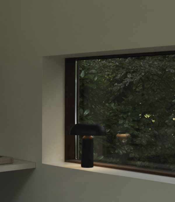 En stilfuld sort NORMANN COPENHAGEN - Porta Bordlampe sidder i en vindueskarm og oplyser et moderne interiør. Vinduet giver udsigt til tæt grønt udenfor, hvilket skaber en fredelig kontrast.