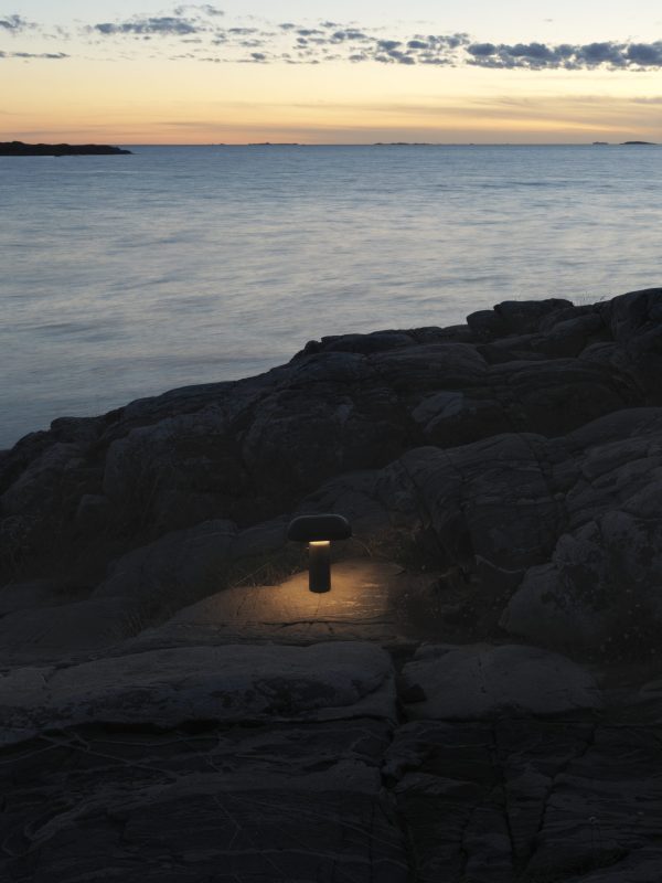 En lille, oplyst NORMANN COPENHAGEN - Porta Bordlampe på en klippefyldt kystlinje i tusmørket, med stille vand og en horisont oplyst af solnedgangens falmende lys.