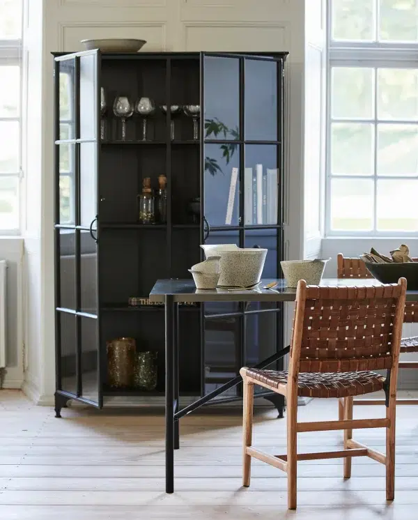 Et rummeligt værelse med en stor sort reol fyldt med bøger og pynteting, et sort skrivebord med en grå skål og en træstol med designmøbler siddepladser ved siden af et vindue med udsigt til haven.