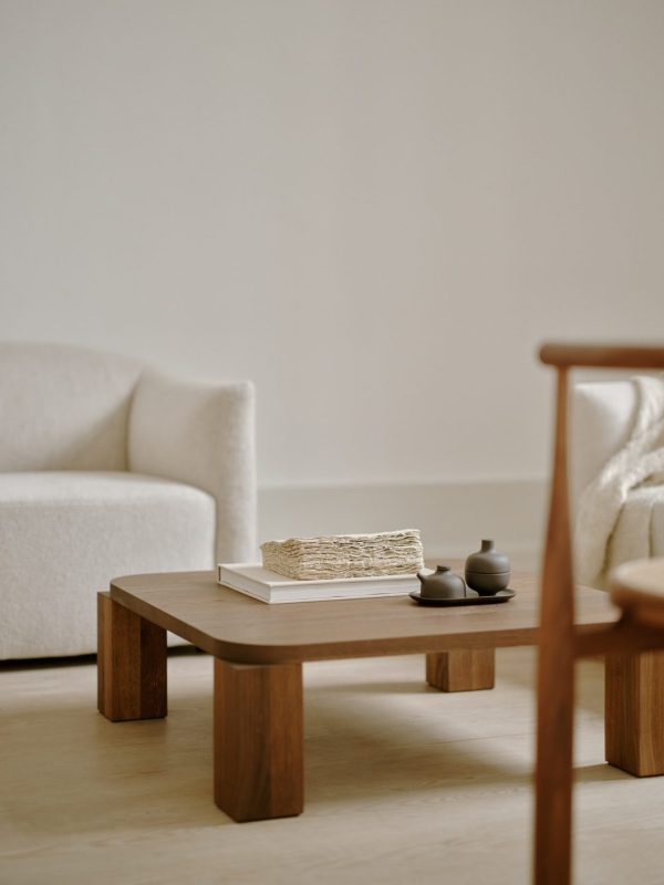 En minimalistisk stue med et Atlas Sofabord 82x82 med en stak bøger og keramiske tekander, flankeret af to lænestole med hvide puder på en almindelig beige baggrund.