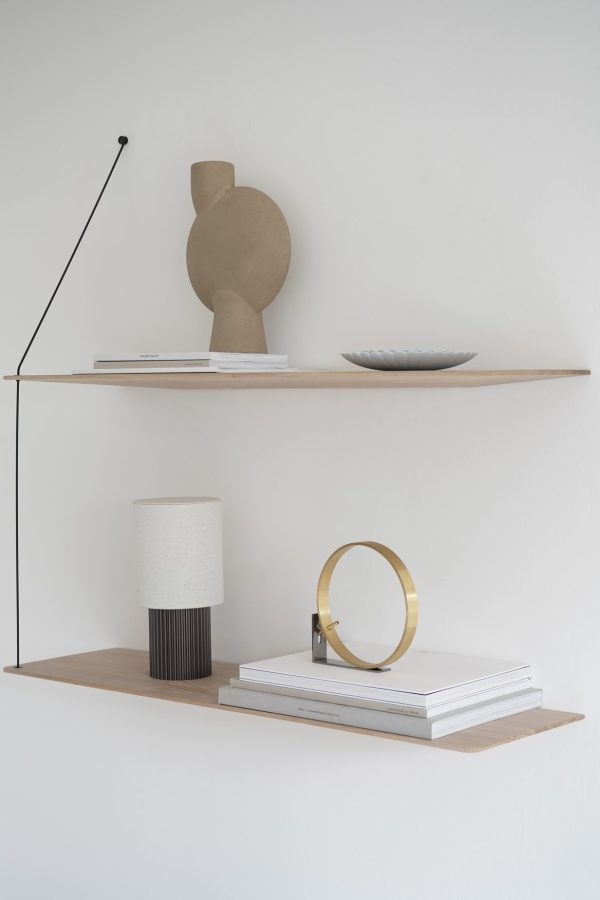 To minimalistiske hylder på en hvid væg med dekorative ting. Den øverste hylde har en skulptur og et fad; den nederste hylde har designmøbler inklusiv en lampe, et rundt spejl og en stak bøger.