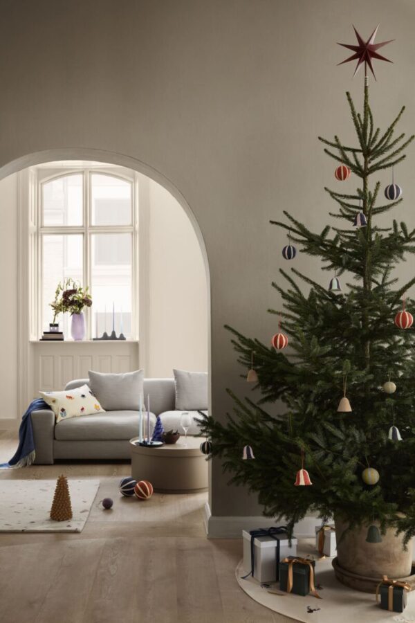 Et hyggeligt julepyntet rum med et stort, ornamenteret træ i forgrunden og et vindue med hvid BROSTE COPENHAGEN - 'Bay' Sofa 3-Personers og puder i baggrunden. blød belysning og gaver på gulvet bidrager til den festlige stemning.