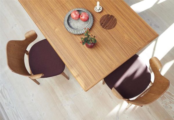 Et spisebord i træ med designstole, en skål med æbler, en lille potteplante og et lys på bordet ovenfra. Gulvet er lyst hårdttræ.