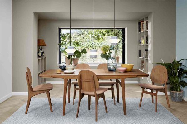 En moderne spisestue med designmøbler, inklusiv et træbord og fire stole sat på et gråt tæppe. En hylde fyldt med bøger, to potteplanter og store vinduer med udsigt over grønne områder er synlige. Afdæmpet belysning og en neutral farvepalet skaber en rolig atmosfære.