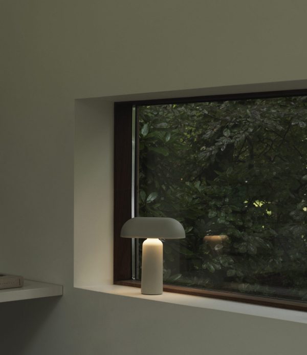 Et svagt oplyst rum med designmøbler og en moderne lampe i vindueskarmen, der oplyser hjørnet. Uden for vinduet er frodigt grønt løv synligt, hvilket tyder på en fredfyldt, naturfyldt udsigt.