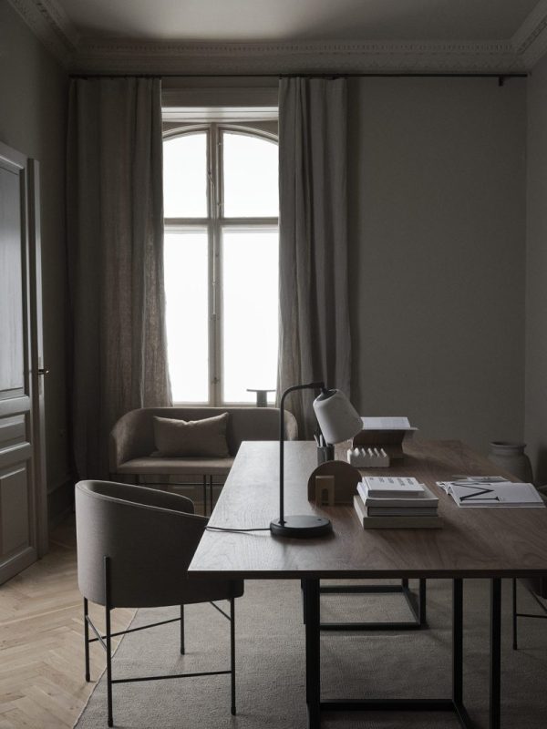 Et svagt oplyst, minimalistisk hjemmekontor med designmøbler inklusive et træbord, en bordlampe, nogle bøger og et par stole, alt sammen sat mod store vinduer draperet med tunge gardiner.