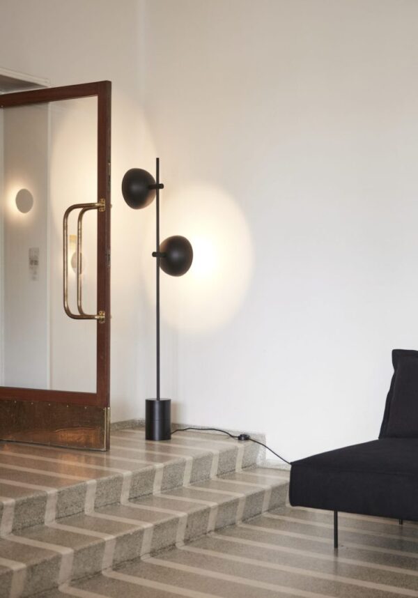 Et stilfuldt interiørdesign med to sfæriske sorte lamper på et stativ, placeret ved siden af en trappe med grå trin og en trædør med glaspaneler og messinghåndtag. En sort sofa er delvist synlig.