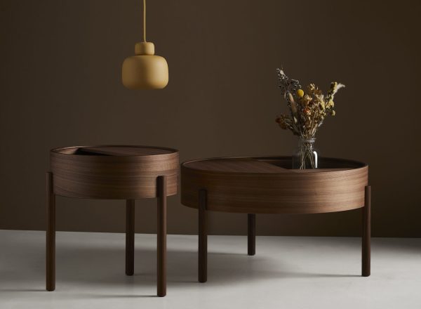 To runde træborde med tynde ben i et rum, det ene med en lille vase med tørrede blomster. En enkelt guldfarvet pendel hænger ovenover i et blødt oplyst interiørdesign.