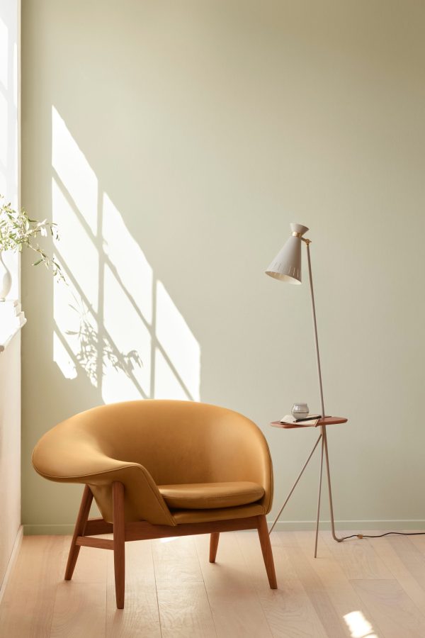 Et minimalistisk interiør med en solbrun moderne lænestol og en slank gulvlampe ved siden af et lille sidebord med en kop, under naturligt lys, der kaster skyggemønstre på en lysegrøn væg.