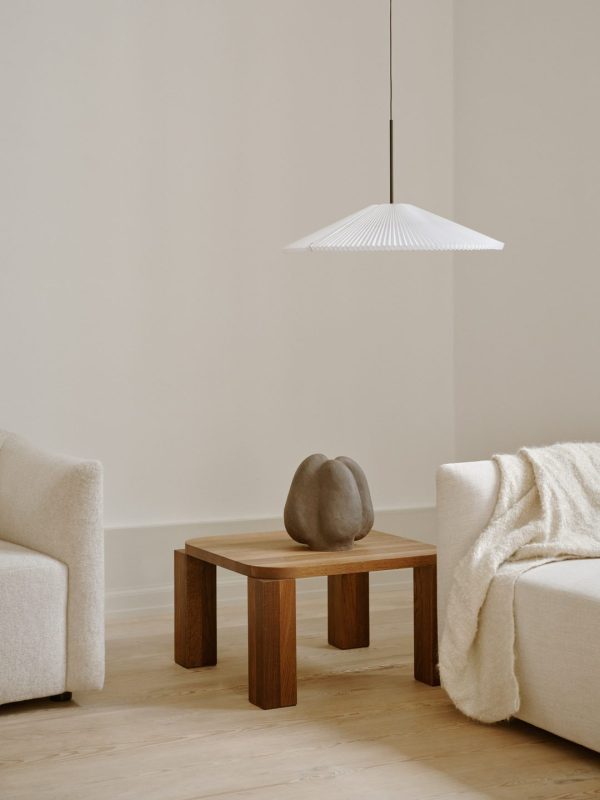 En minimalistisk stue med et NYT VÆRK - Atlas Sofabord 60x60 på et firkantet træbord, flankeret af creme sofaer. over bordet hænger en hvid pendel. lyse trægulve og neutrale vægge er synlige.