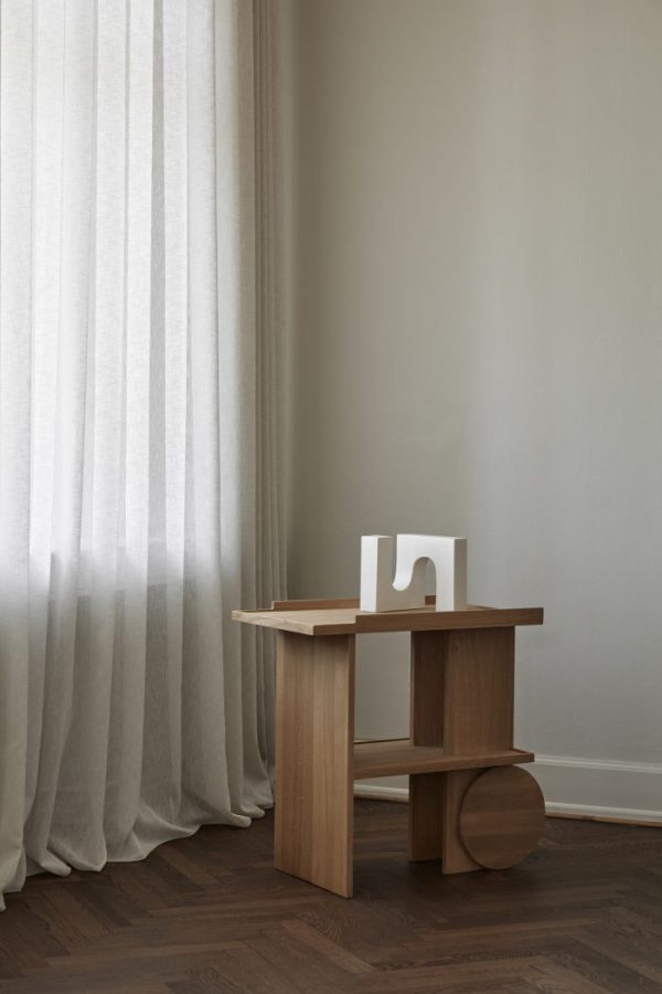 EN KRISTINA DAM - Axis Sidebord står op ad en hvid væg og ved siden af et gennemsigtigt, hvidt gardin. Bordet har en abstrakt hvid skulptur på toppen og har enkle, geometriske designelementer, herunder en stor cirkulær base på den ene side. Gulvet er mørkt hårdttræ.