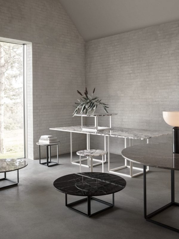 Interiør i et moderne værelse med en række elegante møbler, herunder sideborde i marmor, en skrivebordsopsætning med en plante og en minimalistisk lampe, alt sammen på baggrund af hvide murstensvægge og et stort vindue.