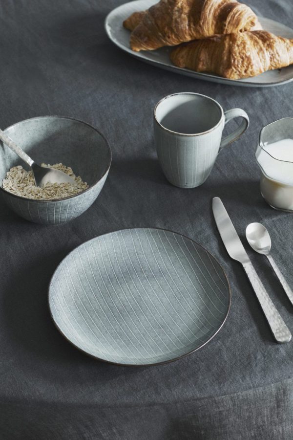 En morgenbordsdækning med en tom stribet keramisk tallerken, en skål med havre, en kande og kop og to croissanter på en tallerken, alt sammen anbragt på en mørkegrå BROSTE COPENHAGEN - 'Nordic Sea' Krus.
