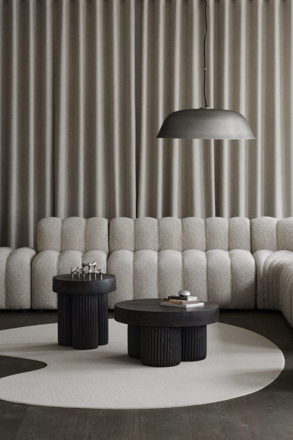 En minimalistisk stue med en lang, beige sofa med bølget tekstur mod en gardinvæg, to sorte cylindriske sofaborde og et stort kuppellys over hovedet. neutral-tonet indretning med et enkelt rundt tæppe på gulvet med udvalgte designmøbler-elementer.