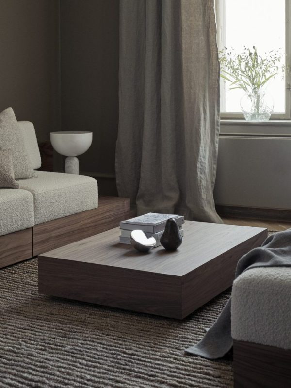 En moderne stue med et lavt, firkantet sofabord i træ med bøger og dekorative genstande på, plys creme sofaer designet af designmøbler og et vindue draperet med tunge gardiner.