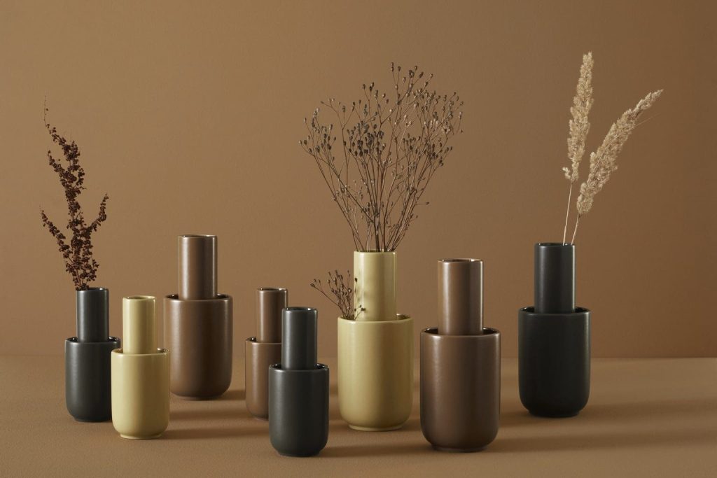 Forskellige keramiske vaser i mat sort, guld og beige arrangeret i et symmetrisk mønster på en beige baggrund, som hver indeholder forskellige typer tørrede planteekstrakter.