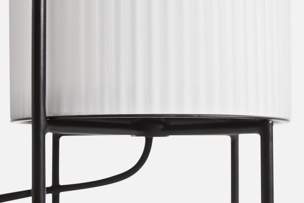 Nærbillede af en WOUD - Ghost Bordlampe med en cylindrisk hvid plisseret skærm monteret på en slank sort metalramme, der inkarnerer morgendagens designæstetik. Fokus er på, hvor skyggen møder rammen.