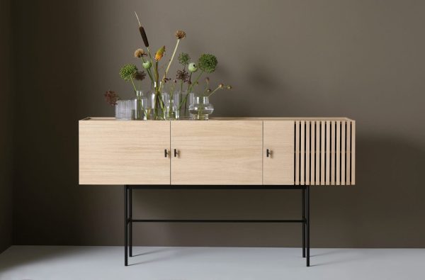 En moderne skænk i lyst træ på en sort metalramme placeret mod en grå baggrund, dekoreret med forskellige glasvaser med grønne og brune tørrede blomster, eksemplificerer Tomorrowsdesigns etos i
